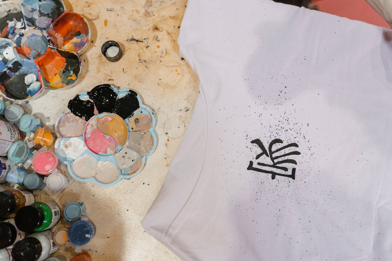 The Art of Drawing Readers In: Your attractive post title goes here Top view of a t-shirt with artistic design surrounded by colorful paint supplies.